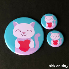 A collection of large and small buttons featuring a smiling purple cat holding a red paper heart. A cute original design by Sick On Sin available on pins, magnets, keychains, etc.